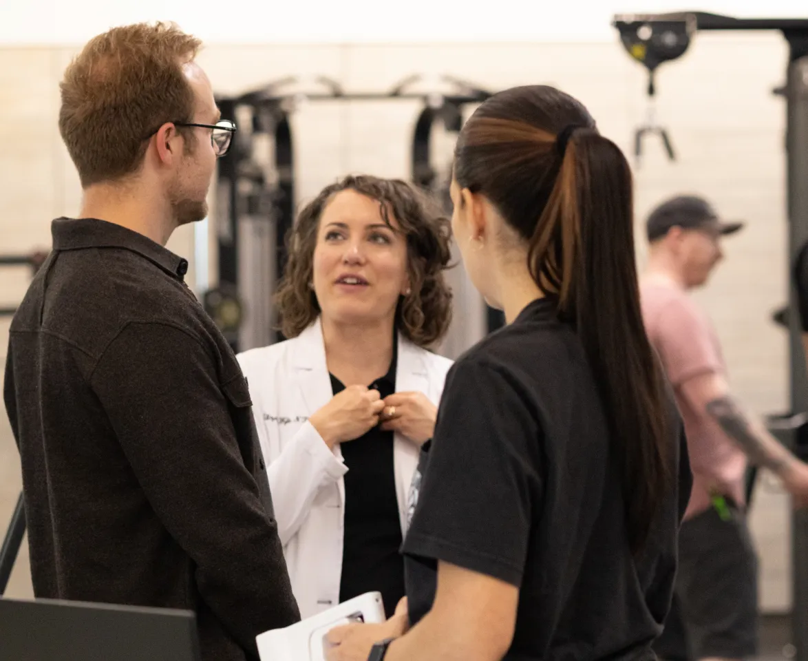 A clinician in a white coat talking with two people in a gym