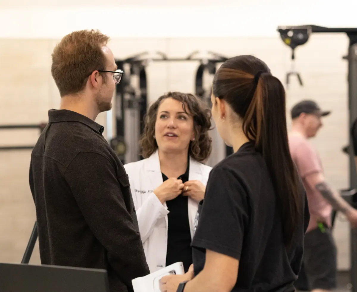 A PRIME Clinics clinician in a white coat talking with two people on a gym floor