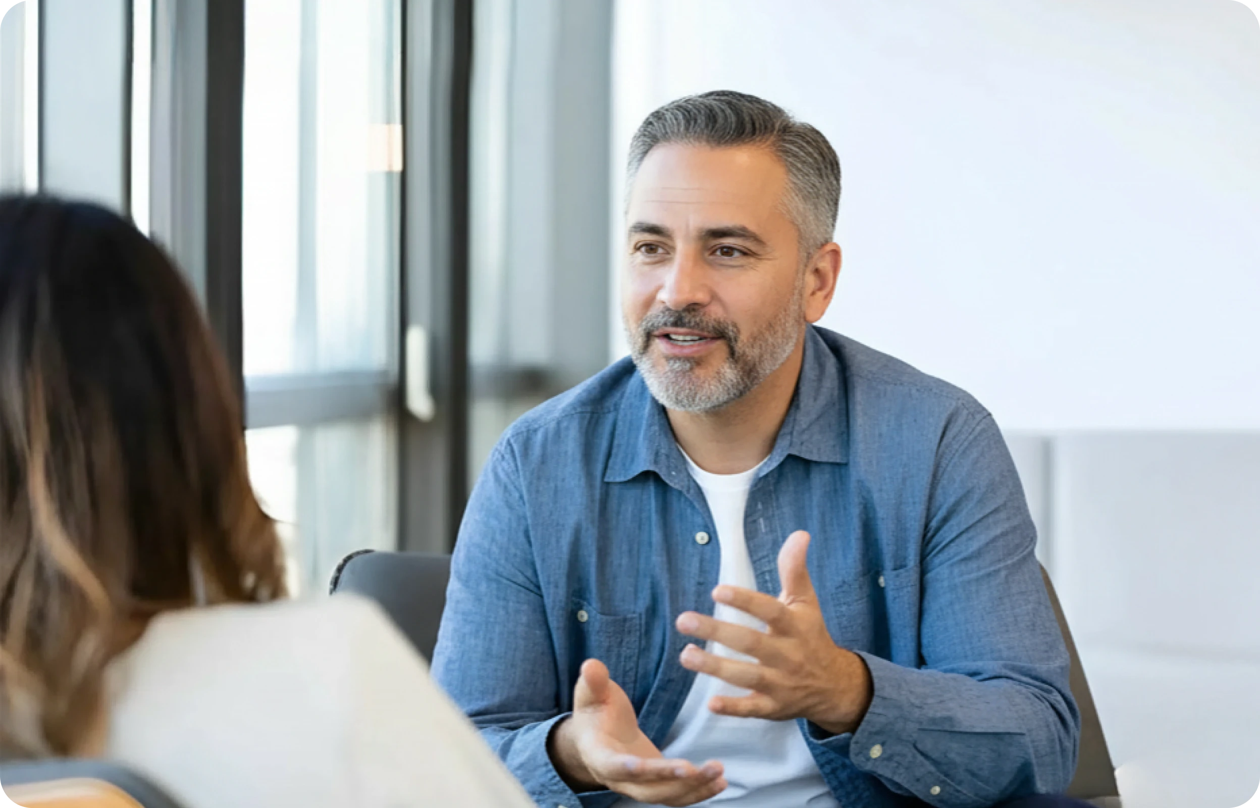 A man with grey hair and a beard, in a blue denim shirt, talking with someone seated across from him