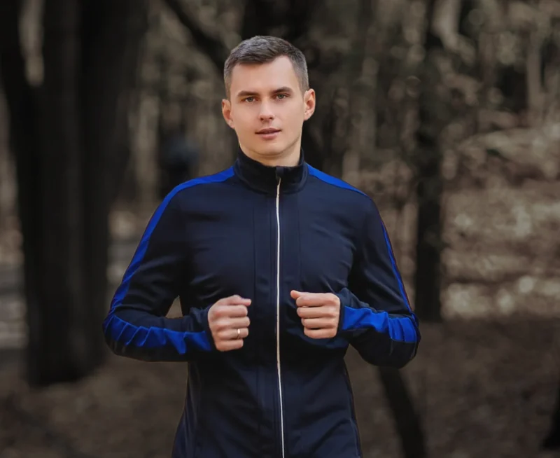 A man in a navy and blue track jacket running on a wooded path